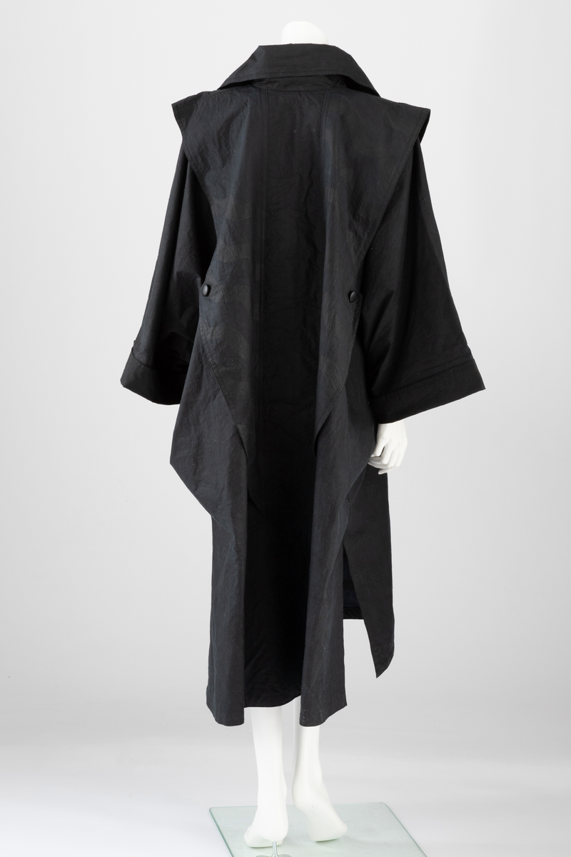 Svart regnkappe i kimonofasong med oppbrett på ermene og stor, rett, utbrettet krage. Kappen er fôret med svart, tynt bomullsstoff. Løst stoffstykke foran som går ned i en spiss, sydd til stolpen og festet med en knapp i ytterkant. Bak er det løse stykket sydd ned til en søm 5,5cm fra midt bak på begge sider av midten. Kappen lukkes med trykknapper og en knapp og knapphull ved halsen. Rygg og forstykke er sydd i ett stykke. Krage, oppbrett på ermene og stoffstykket foran er ensfarget svart. Stykket bak har bølgeformet dekor (stofftrykk). Tilhørende ensfarget svart, sirkelrund, flat lue. Oppå pullen er det sydd fast to løse, doble kanter, festet med en knapp i yttersiden.