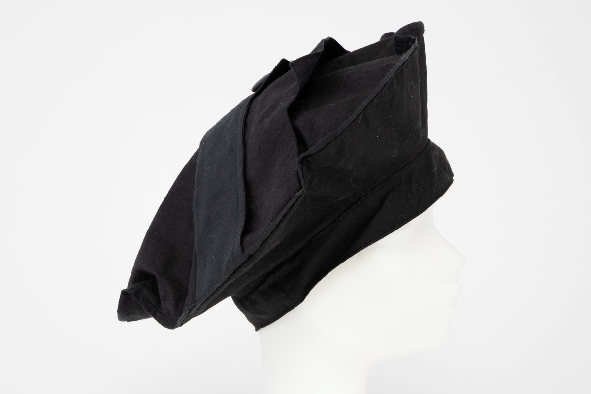 Svart regnkappe i kimonofasong med oppbrett på ermene og stor, rett, utbrettet krage. Kappen er fôret med svart, tynt bomullsstoff. Løst stoffstykke foran som går ned i en spiss, sydd til stolpen og festet med en knapp i ytterkant. Bak er det løse stykket sydd ned til en søm 5,5cm fra midt bak på begge sider av midten. Kappen lukkes med trykknapper og en knapp og knapphull ved halsen. Rygg og forstykke er sydd i ett stykke. Krage, oppbrett på ermene og stoffstykket foran er ensfarget svart. Stykket bak har bølgeformet dekor (stofftrykk). Tilhørende ensfarget svart, sirkelrund, flat lue. Oppå pullen er det sydd fast to løse, doble kanter, festet med en knapp i yttersiden.