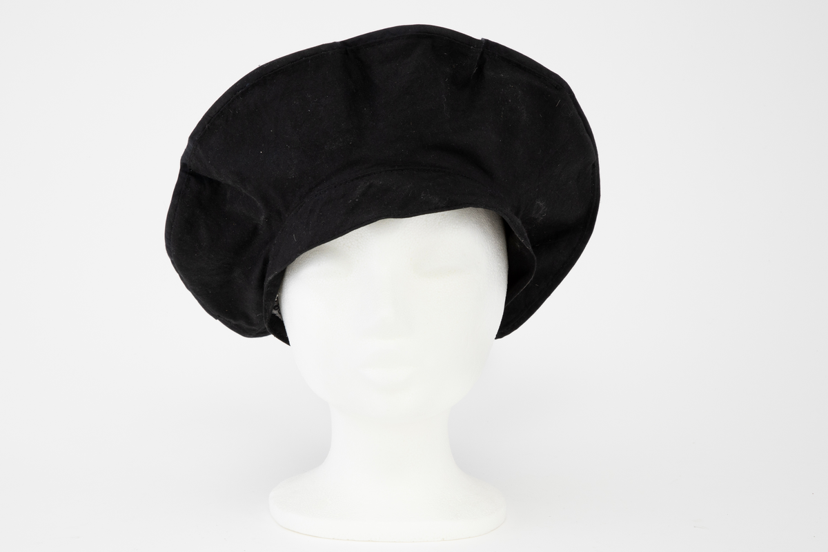 Svart regnkappe i kimonofasong med oppbrett på ermene og stor, rett, utbrettet krage. Kappen er fôret med svart, tynt bomullsstoff. Løst stoffstykke foran som går ned i en spiss, sydd til stolpen og festet med en knapp i ytterkant. Bak er det løse stykket sydd ned til en søm 5,5cm fra midt bak på begge sider av midten. Kappen lukkes med trykknapper og en knapp og knapphull ved halsen. Rygg og forstykke er sydd i ett stykke. Krage, oppbrett på ermene og stoffstykket foran er ensfarget svart. Stykket bak har bølgeformet dekor (stofftrykk). Tilhørende ensfarget svart, sirkelrund, flat lue. Oppå pullen er det sydd fast to løse, doble kanter, festet med en knapp i yttersiden.