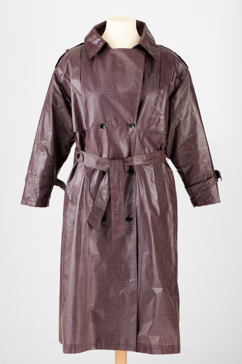 Pepitarutet regnkåpe i rosa, lilla og grønn. Kåpen er dobbelspent og har utenpåsatte bærestykker foran og bak. Bak er bærestykket delt i to med en motfold med knapp og knapphull midt bak. Kåpen har stor slag og krage. Stropper med knapphull og knapp over skuldrene og på ermene. Skrå stikklomme i hver side. Knyttebelte med hempe og knapp i livet.