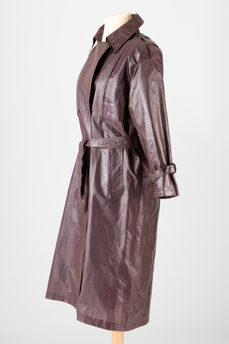 Pepitarutet regnkåpe i rosa, lilla og grønn. Kåpen er dobbelspent og har utenpåsatte bærestykker foran og bak. Bak er bærestykket delt i to med en motfold med knapp og knapphull midt bak. Kåpen har stor slag og krage. Stropper med knapphull og knapp over skuldrene og på ermene. Skrå stikklomme i hver side. Knyttebelte med hempe og knapp i livet.