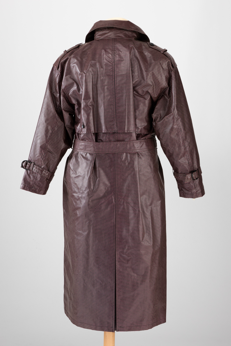 Pepitarutet regnkåpe i rosa, lilla og grønn. Kåpen er dobbelspent og har utenpåsatte bærestykker foran og bak. Bak er bærestykket delt i to med en motfold med knapp og knapphull midt bak. Kåpen har stor slag og krage. Stropper med knapphull og knapp over skuldrene og på ermene. Skrå stikklomme i hver side. Knyttebelte med hempe og knapp i livet.