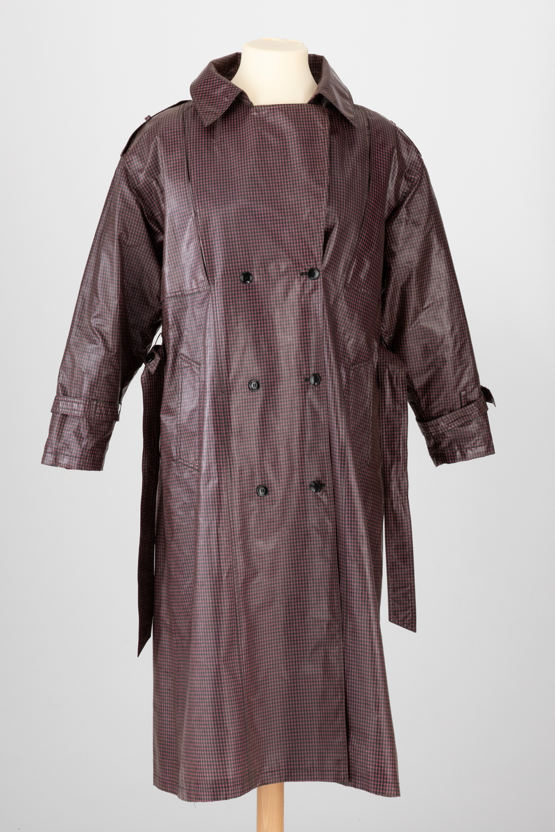 Pepitarutet regnkåpe i rosa, lilla og grønn. Kåpen er dobbelspent og har utenpåsatte bærestykker foran og bak. Bak er bærestykket delt i to med en motfold med knapp og knapphull midt bak. Kåpen har stor slag og krage. Stropper med knapphull og knapp over skuldrene og på ermene. Skrå stikklomme i hver side. Knyttebelte med hempe og knapp i livet.