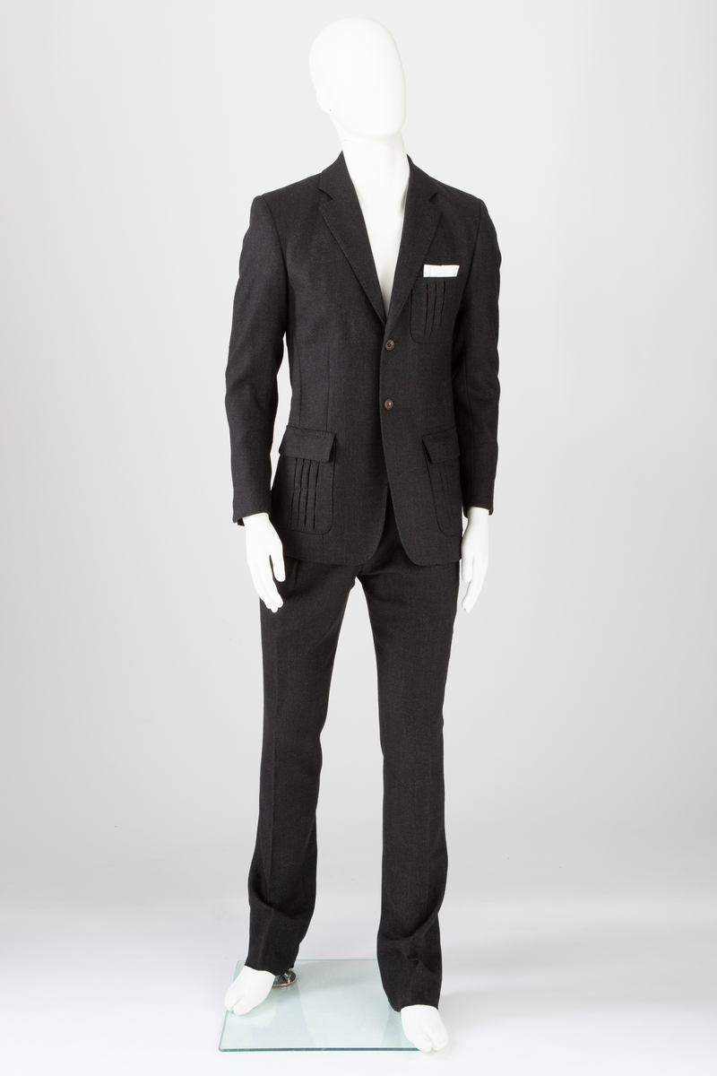 Mørk grå ulldress sydd av fast materiale i toskaftsbinding. Jakke og bukse, levert med matchende tversoversløyfe i samme materiale.
Jakken er figursydd og enkeltkneppet, har avrundede forstykker og lukkes med to knapper. Den er konstruert av to bakstykker med midtsøm og panelsømmer, to sidestykker under ermene og to forstykker. Panelsømmene på ryggen er stukket ved siden av sømmen. De ender i hver sin splitt nederst. Tosøms-ermer med splitt nederst. Påsydde lommer nede på hver forbredd og venstre bryst. Jakken er delvis fôret med blankt Polyester-/Viskosemateriale, mørk fiolett prydet med påtrykte strektegninger av verktøy i lilla. Også alle ytterkanter av ull- og fôrstoff er kantet med lilla skråbånd.
Buksen har gylf lukket med tre knapper og hekte pluss knapp i linningen. Skråstilte stikklommer og et lite legg på hver side av gylfen og på hver side, over baken. Venstre forstykke har også en kraftig, blank, dekorativ glidelås innsatt oppe ved linningen. Denne kan ikke åpnes og er kun dekorativ. Åpen lomme øverst på høyre bakstykke med påsydd, dekorativt belegg i tre-takket kronefasong i overkant. Bena er kun kantet med sikksakk-søm nederst. Fôret ned foran, til og med knærne med fôrsilke. Lommene er fôret med bomullsmateriale. Klokkelomme nedenfor linningen foran på høyre fremstykke.