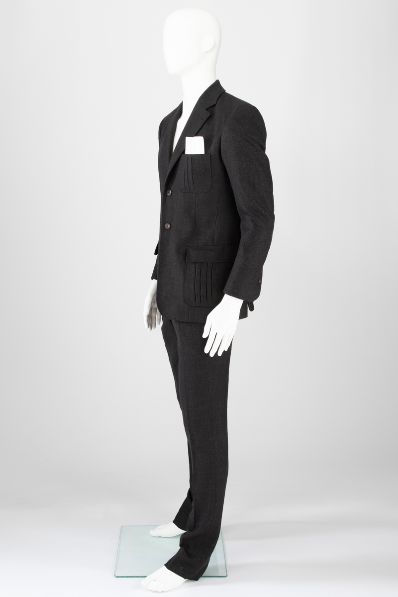 Mørk grå ulldress sydd av fast materiale i toskaftsbinding. Jakke og bukse, levert med matchende tversoversløyfe i samme materiale.
Jakken er figursydd og enkeltkneppet, har avrundede forstykker og lukkes med to knapper. Den er konstruert av to bakstykker med midtsøm og panelsømmer, to sidestykker under ermene og to forstykker. Panelsømmene på ryggen er stukket ved siden av sømmen. De ender i hver sin splitt nederst. Tosøms-ermer med splitt nederst. Påsydde lommer nede på hver forbredd og venstre bryst. Jakken er delvis fôret med blankt Polyester-/Viskosemateriale, mørk fiolett prydet med påtrykte strektegninger av verktøy i lilla. Også alle ytterkanter av ull- og fôrstoff er kantet med lilla skråbånd.
Buksen har gylf lukket med tre knapper og hekte pluss knapp i linningen. Skråstilte stikklommer og et lite legg på hver side av gylfen og på hver side, over baken. Venstre forstykke har også en kraftig, blank, dekorativ glidelås innsatt oppe ved linningen. Denne kan ikke åpnes og er kun dekorativ. Åpen lomme øverst på høyre bakstykke med påsydd, dekorativt belegg i tre-takket kronefasong i overkant. Bena er kun kantet med sikksakk-søm nederst. Fôret ned foran, til og med knærne med fôrsilke. Lommene er fôret med bomullsmateriale. Klokkelomme nedenfor linningen foran på høyre fremstykke.