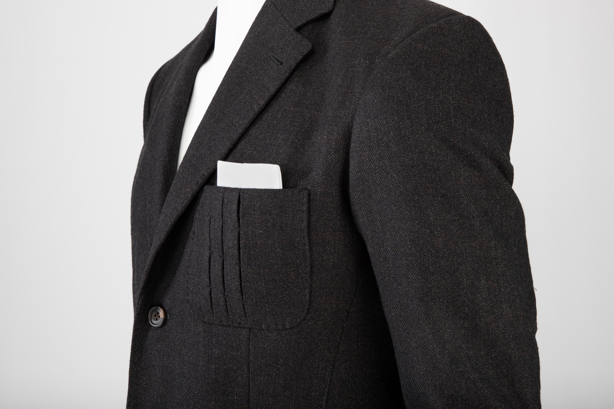 Mørk grå ulldress sydd av fast materiale i toskaftsbinding. Jakke og bukse, levert med matchende tversoversløyfe i samme materiale.
Jakken er figursydd og enkeltkneppet, har avrundede forstykker og lukkes med to knapper. Den er konstruert av to bakstykker med midtsøm og panelsømmer, to sidestykker under ermene og to forstykker. Panelsømmene på ryggen er stukket ved siden av sømmen. De ender i hver sin splitt nederst. Tosøms-ermer med splitt nederst. Påsydde lommer nede på hver forbredd og venstre bryst. Jakken er delvis fôret med blankt Polyester-/Viskosemateriale, mørk fiolett prydet med påtrykte strektegninger av verktøy i lilla. Også alle ytterkanter av ull- og fôrstoff er kantet med lilla skråbånd.
Buksen har gylf lukket med tre knapper og hekte pluss knapp i linningen. Skråstilte stikklommer og et lite legg på hver side av gylfen og på hver side, over baken. Venstre forstykke har også en kraftig, blank, dekorativ glidelås innsatt oppe ved linningen. Denne kan ikke åpnes og er kun dekorativ. Åpen lomme øverst på høyre bakstykke med påsydd, dekorativt belegg i tre-takket kronefasong i overkant. Bena er kun kantet med sikksakk-søm nederst. Fôret ned foran, til og med knærne med fôrsilke. Lommene er fôret med bomullsmateriale. Klokkelomme nedenfor linningen foran på høyre fremstykke.