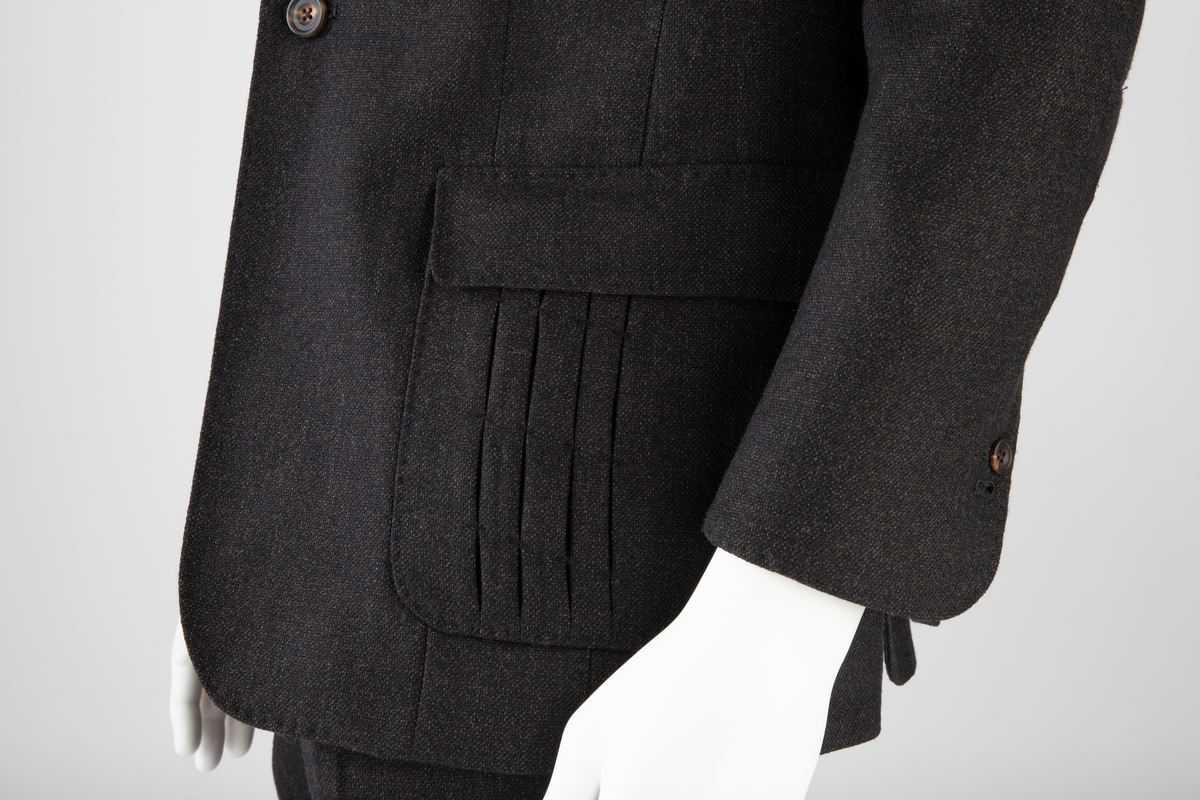 Mørk grå ulldress sydd av fast materiale i toskaftsbinding. Jakke og bukse, levert med matchende tversoversløyfe i samme materiale.
Jakken er figursydd og enkeltkneppet, har avrundede forstykker og lukkes med to knapper. Den er konstruert av to bakstykker med midtsøm og panelsømmer, to sidestykker under ermene og to forstykker. Panelsømmene på ryggen er stukket ved siden av sømmen. De ender i hver sin splitt nederst. Tosøms-ermer med splitt nederst. Påsydde lommer nede på hver forbredd og venstre bryst. Jakken er delvis fôret med blankt Polyester-/Viskosemateriale, mørk fiolett prydet med påtrykte strektegninger av verktøy i lilla. Også alle ytterkanter av ull- og fôrstoff er kantet med lilla skråbånd.
Buksen har gylf lukket med tre knapper og hekte pluss knapp i linningen. Skråstilte stikklommer og et lite legg på hver side av gylfen og på hver side, over baken. Venstre forstykke har også en kraftig, blank, dekorativ glidelås innsatt oppe ved linningen. Denne kan ikke åpnes og er kun dekorativ. Åpen lomme øverst på høyre bakstykke med påsydd, dekorativt belegg i tre-takket kronefasong i overkant. Bena er kun kantet med sikksakk-søm nederst. Fôret ned foran, til og med knærne med fôrsilke. Lommene er fôret med bomullsmateriale. Klokkelomme nedenfor linningen foran på høyre fremstykke.