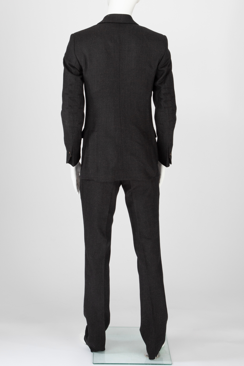 Mørk grå ulldress sydd av fast materiale i toskaftsbinding. Jakke og bukse, levert med matchende tversoversløyfe i samme materiale.
Jakken er figursydd og enkeltkneppet, har avrundede forstykker og lukkes med to knapper. Den er konstruert av to bakstykker med midtsøm og panelsømmer, to sidestykker under ermene og to forstykker. Panelsømmene på ryggen er stukket ved siden av sømmen. De ender i hver sin splitt nederst. Tosøms-ermer med splitt nederst. Påsydde lommer nede på hver forbredd og venstre bryst. Jakken er delvis fôret med blankt Polyester-/Viskosemateriale, mørk fiolett prydet med påtrykte strektegninger av verktøy i lilla. Også alle ytterkanter av ull- og fôrstoff er kantet med lilla skråbånd.
Buksen har gylf lukket med tre knapper og hekte pluss knapp i linningen. Skråstilte stikklommer og et lite legg på hver side av gylfen og på hver side, over baken. Venstre forstykke har også en kraftig, blank, dekorativ glidelås innsatt oppe ved linningen. Denne kan ikke åpnes og er kun dekorativ. Åpen lomme øverst på høyre bakstykke med påsydd, dekorativt belegg i tre-takket kronefasong i overkant. Bena er kun kantet med sikksakk-søm nederst. Fôret ned foran, til og med knærne med fôrsilke. Lommene er fôret med bomullsmateriale. Klokkelomme nedenfor linningen foran på høyre fremstykke.