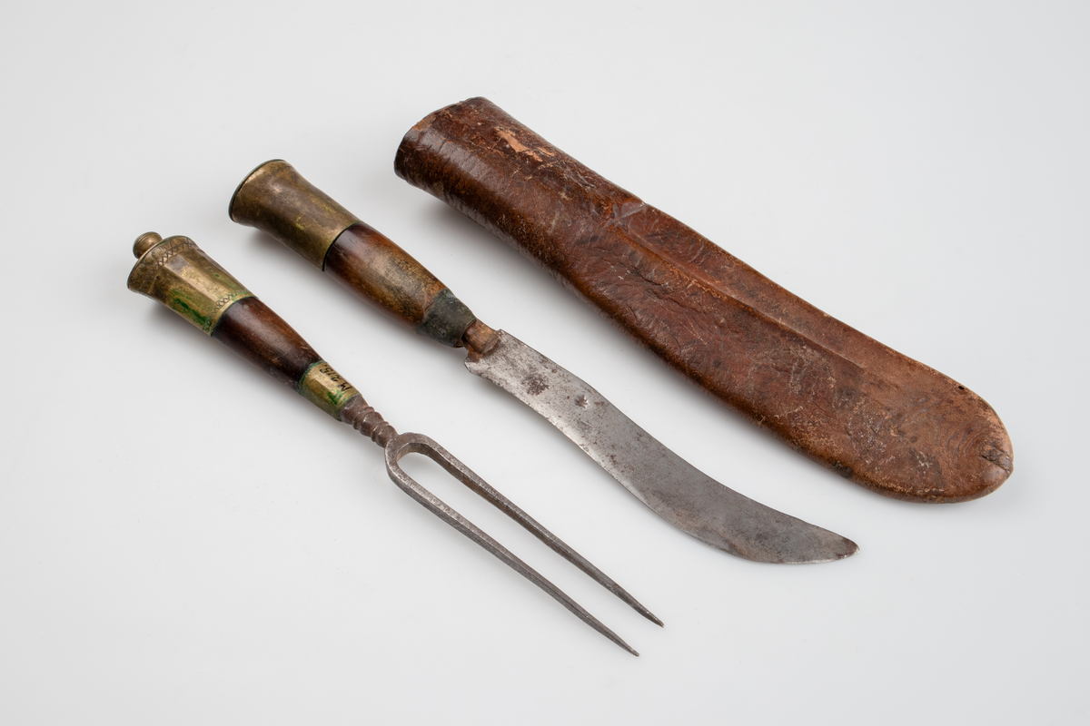 Kniv och gaffel, i läderslida. Gaffel med två klor av metall. Rundat, något koniskt, handtag av trä och mässing. Handtaget avslutas med en rund knopp, även den av mässing. Kniv med svängt blad, spetsigt framtill. Även kniven med rundat, nogåt koniskt, trähandtag med mässingsdetaljer. 
Läderfodral med märkning I 182UUSSDS.

Inskrivet i huvudkatalog 1886.