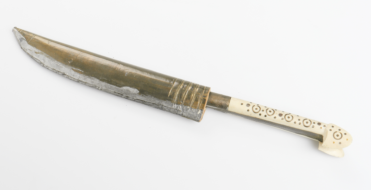 Bulgarisk kniv med handtag av metall och ben. Dekorerat med utsirningar. Fodral av trä med metallöverdrag. 

Inskrivet i huvudbok år 1910.
