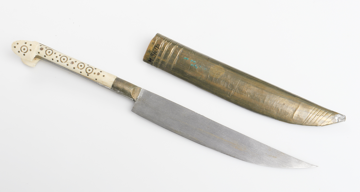 Bulgarisk kniv med handtag av metall och ben. Dekorerat med utsirningar. Fodral av trä med metallöverdrag. 

Inskrivet i huvudbok år 1910.