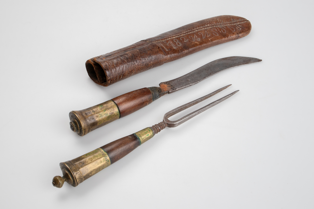Kniv och gaffel, i läderslida. Gaffel med två klor av metall. Rundat, något koniskt, handtag av trä och mässing. Handtaget avslutas med en rund knopp, även den av mässing. Kniv med svängt blad, spetsigt framtill. Även kniven med rundat, nogåt koniskt, trähandtag med mässingsdetaljer. 
Läderfodral med märkning I 182UUSSDS.

Inskrivet i huvudkatalog 1886.