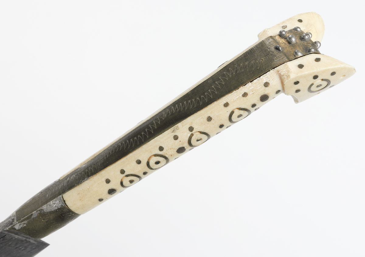 Bulgarisk kniv med handtag av metall och ben. Dekorerat med utsirningar. Fodral av trä med metallöverdrag. 

Inskrivet i huvudbok år 1910.