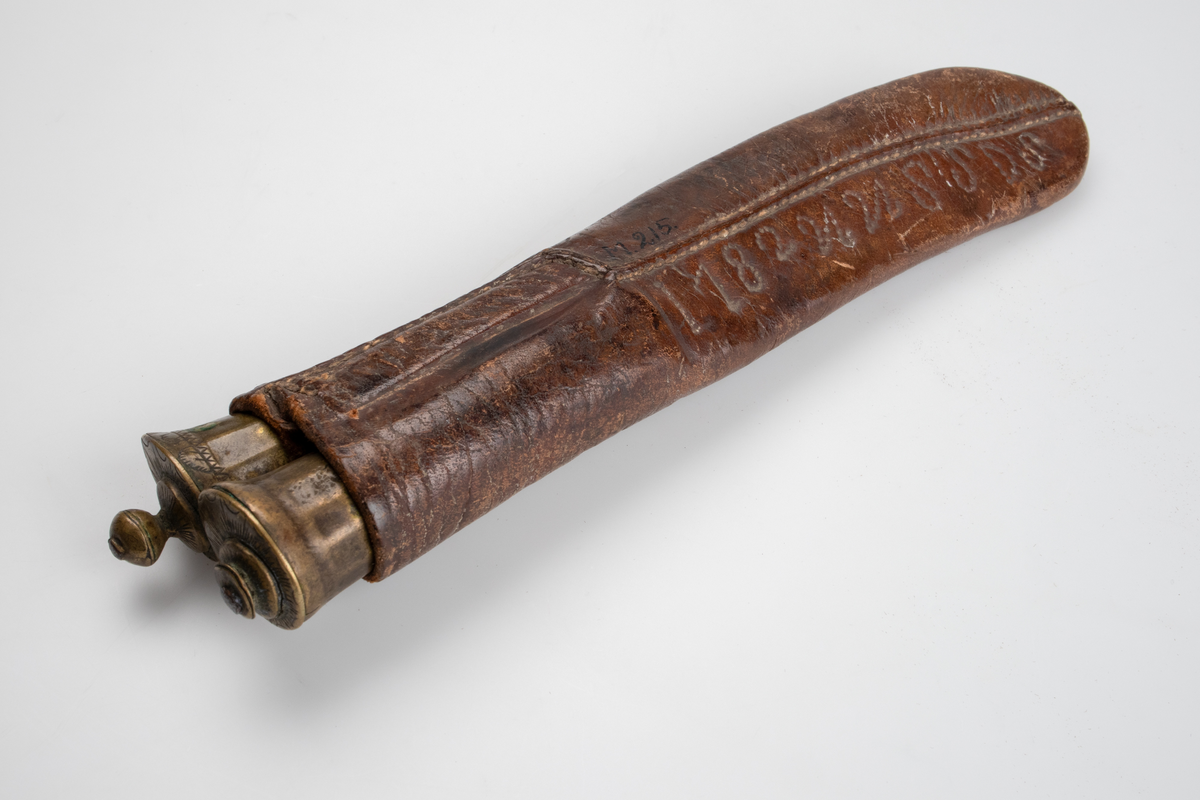 Kniv och gaffel, i läderslida. Gaffel med två klor av metall. Rundat, något koniskt, handtag av trä och mässing. Handtaget avslutas med en rund knopp, även den av mässing. Kniv med svängt blad, spetsigt framtill. Även kniven med rundat, nogåt koniskt, trähandtag med mässingsdetaljer. 
Läderfodral med märkning I 182UUSSDS.

Inskrivet i huvudkatalog 1886.