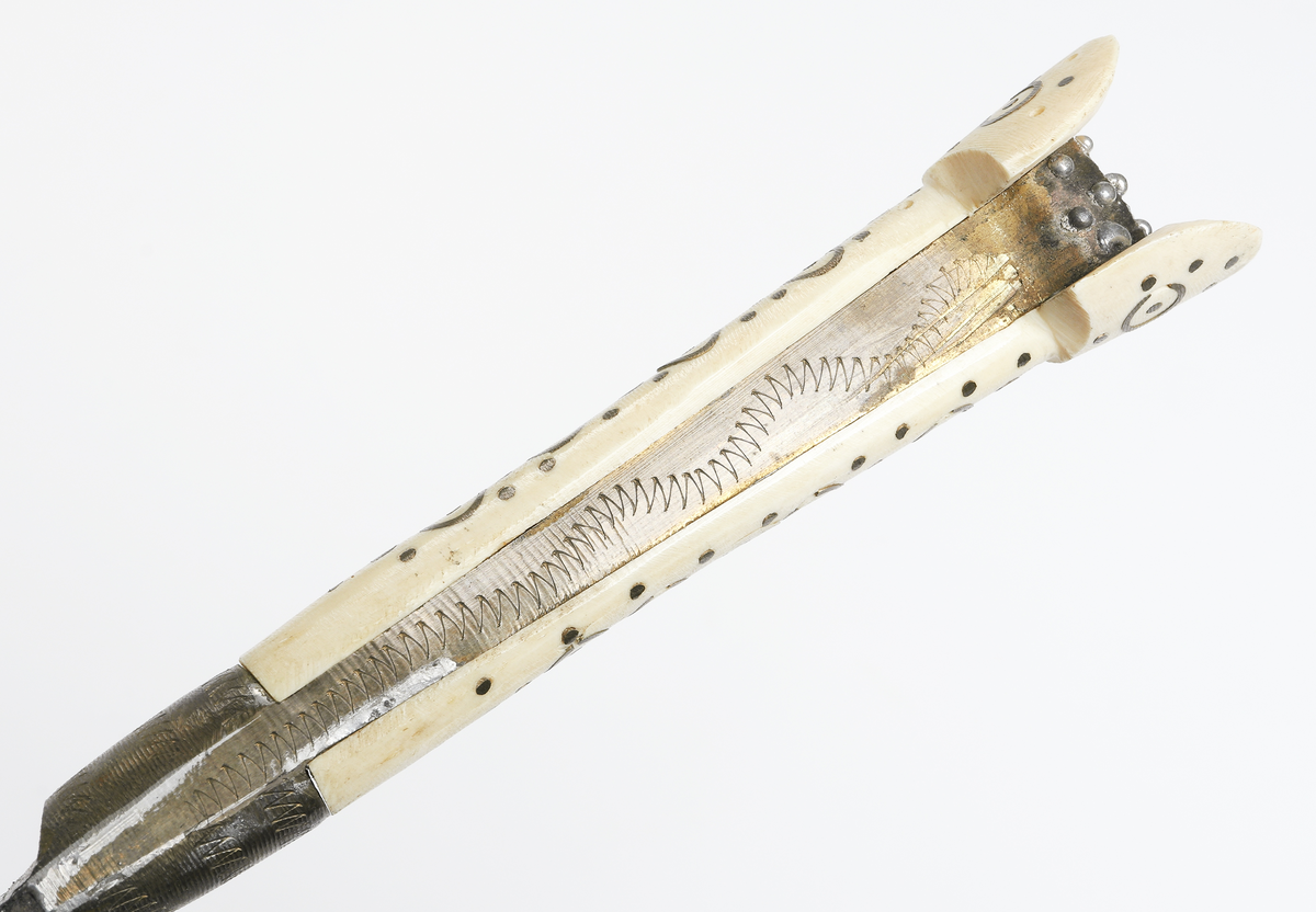 Bulgarisk kniv med handtag av metall och ben. Dekorerat med utsirningar. Fodral av trä med metallöverdrag. 

Inskrivet i huvudbok år 1910.