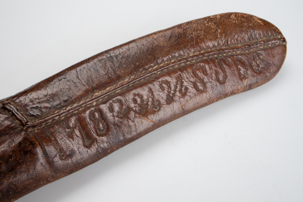 Kniv och gaffel, i läderslida. Gaffel med två klor av metall. Rundat, något koniskt, handtag av trä och mässing. Handtaget avslutas med en rund knopp, även den av mässing. Kniv med svängt blad, spetsigt framtill. Även kniven med rundat, nogåt koniskt, trähandtag med mässingsdetaljer. 
Läderfodral med märkning I 182UUSSDS.

Inskrivet i huvudkatalog 1886.