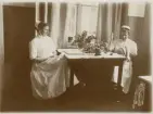 Två sjuksköterskor sitter och syr i ett personalrum. 
Till höger fröken Stina Eksandh (i Sofiasyster-uniform), dotter t. häradshövding Theodor Eksandh och h.h. Anna.