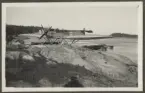 Bilden visar ett sjöflygplan typ Heinkel HE 2 i en skärgårdsmiljö. Två män arbetar framför flygplanet.