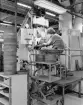 PRESSNING. Denna bild får exemplifiera en icke automatiserad arbetsplats för pressning. Operatören sitter här framför pressen och plockar för hand de pressade ämnena från pressbordet till grafittallriken. Arbetet var ensidigt och monotont och helt styrt av pressen som gick kontinuerligt. Avbrott skedde endast i samband med att vissa kontrollmoment utfördes. En operatör betjänade en press till skillnad mot dagens förhållande där operatören övervakar flera pressar.