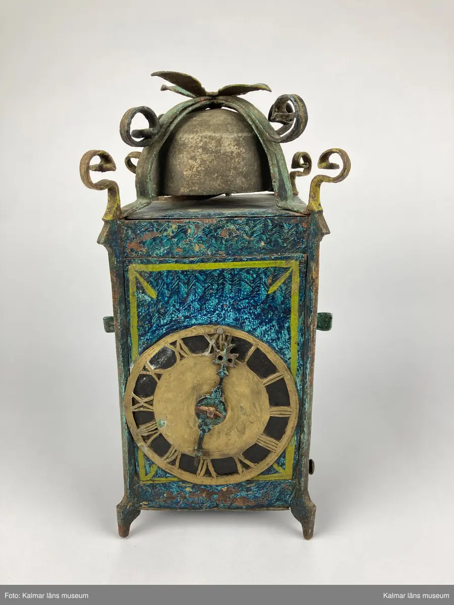 KLM 2798. Bordsur. Rektangulär stomme av järn med öppningsbara sidor. Upptill smidda detaljer samt ringklocka. På ena sidan urtavla av mässing med en visare av järn. Urverk av järn och mässing med släphaksgång. Fodralet är målat blått med gula kantlister, på sidoväggarna en lyra i gult. Uret har fyra fötter. På baksidan ögla av järn. En pendel av metall tillhör. Lodlinor, lod och trissor saknas.