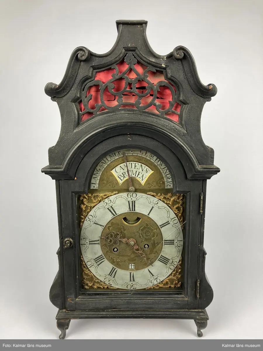 KLM 2268. Bordsur. Fodral av svartlackerat trä med genombrutna ornament med rött (blekt) siden bakom. Profilerat krön. Fodralet är på baksidan försedd med en dörr. Urverk av mässing. Bakom en glasförsedd dörr urtavlan av tenn och mässing med brännförgyllda ornament. Urtavlan är signerad: Castens Bremen. Fodralet har fyra korta profilerade ben.