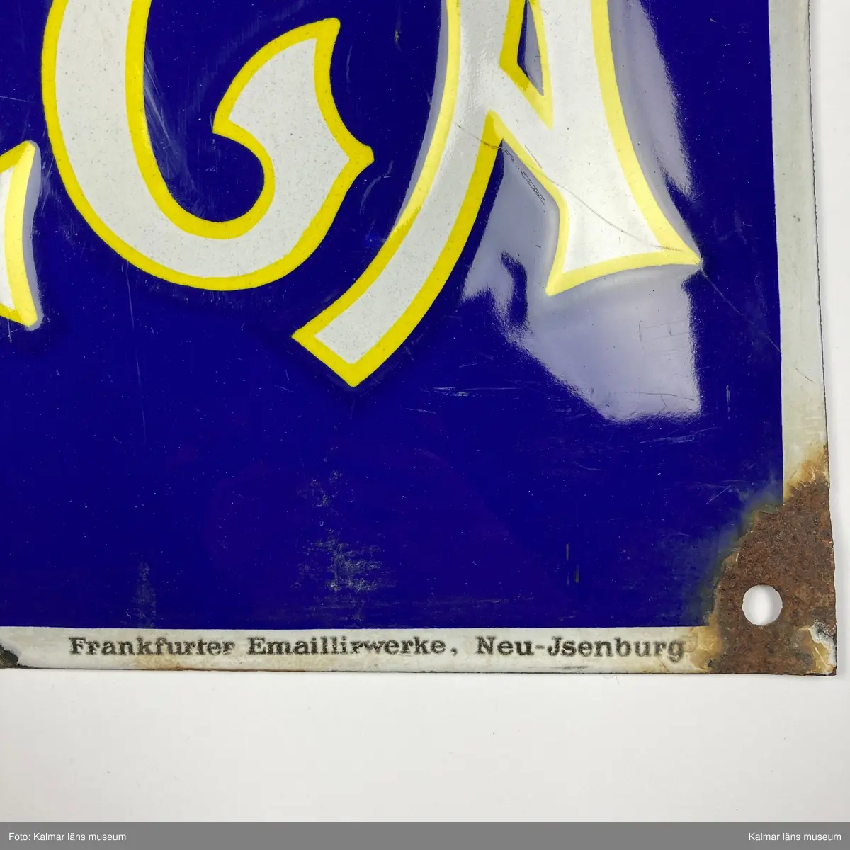 KLM 24993:170. Skylt. Rektangulär och något kupad skylt i emaljerad metall. Blå bakgrund med motiv i gult och vitt föreställande ett fickur och omegatecken samt texten OMEGA. I nedre högra hörnet text: Frankfurter Emaillirwerke, Neu-Jsenburg.