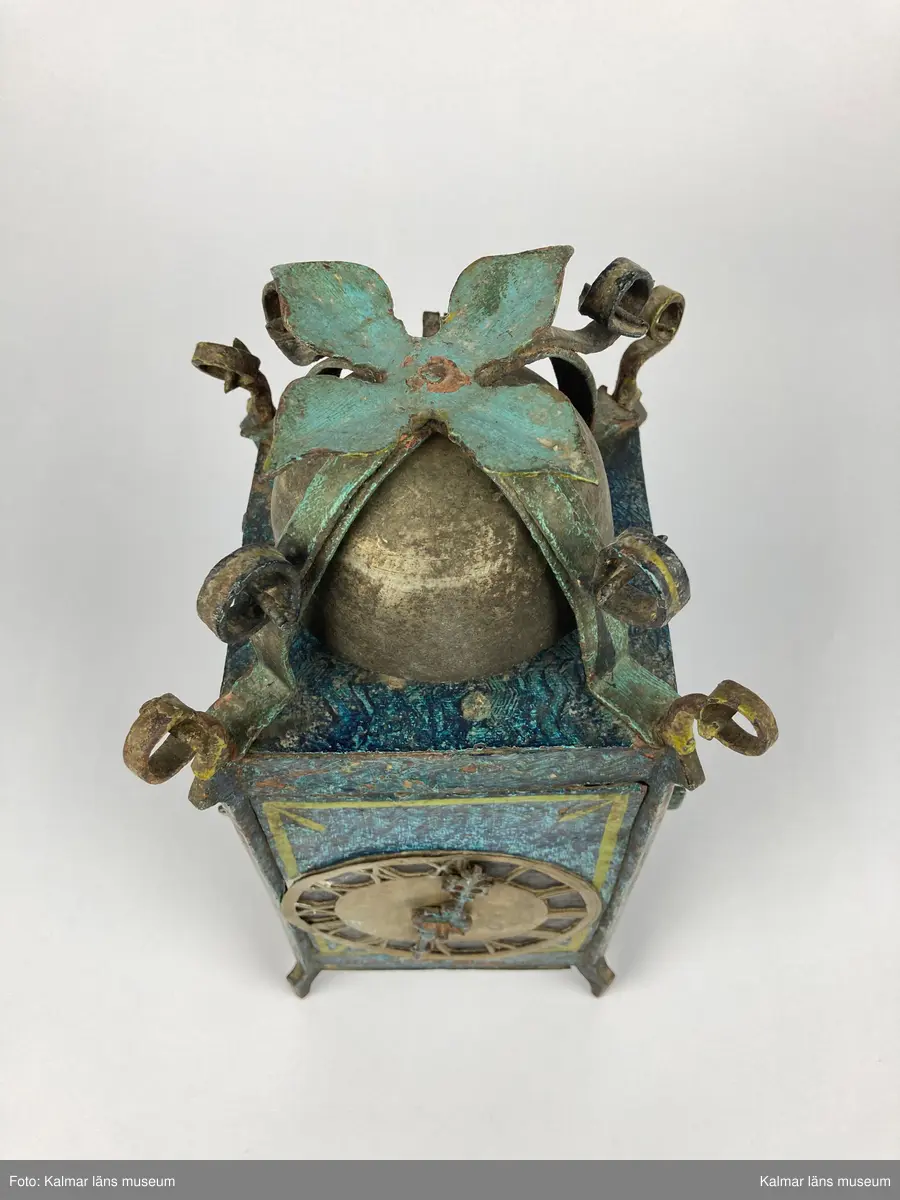 KLM 2798. Bordsur. Rektangulär stomme av järn med öppningsbara sidor. Upptill smidda detaljer samt ringklocka. På ena sidan urtavla av mässing med en visare av järn. Urverk av järn och mässing med släphaksgång. Fodralet är målat blått med gula kantlister, på sidoväggarna en lyra i gult. Uret har fyra fötter. På baksidan ögla av järn. En pendel av metall tillhör. Lodlinor, lod och trissor saknas.