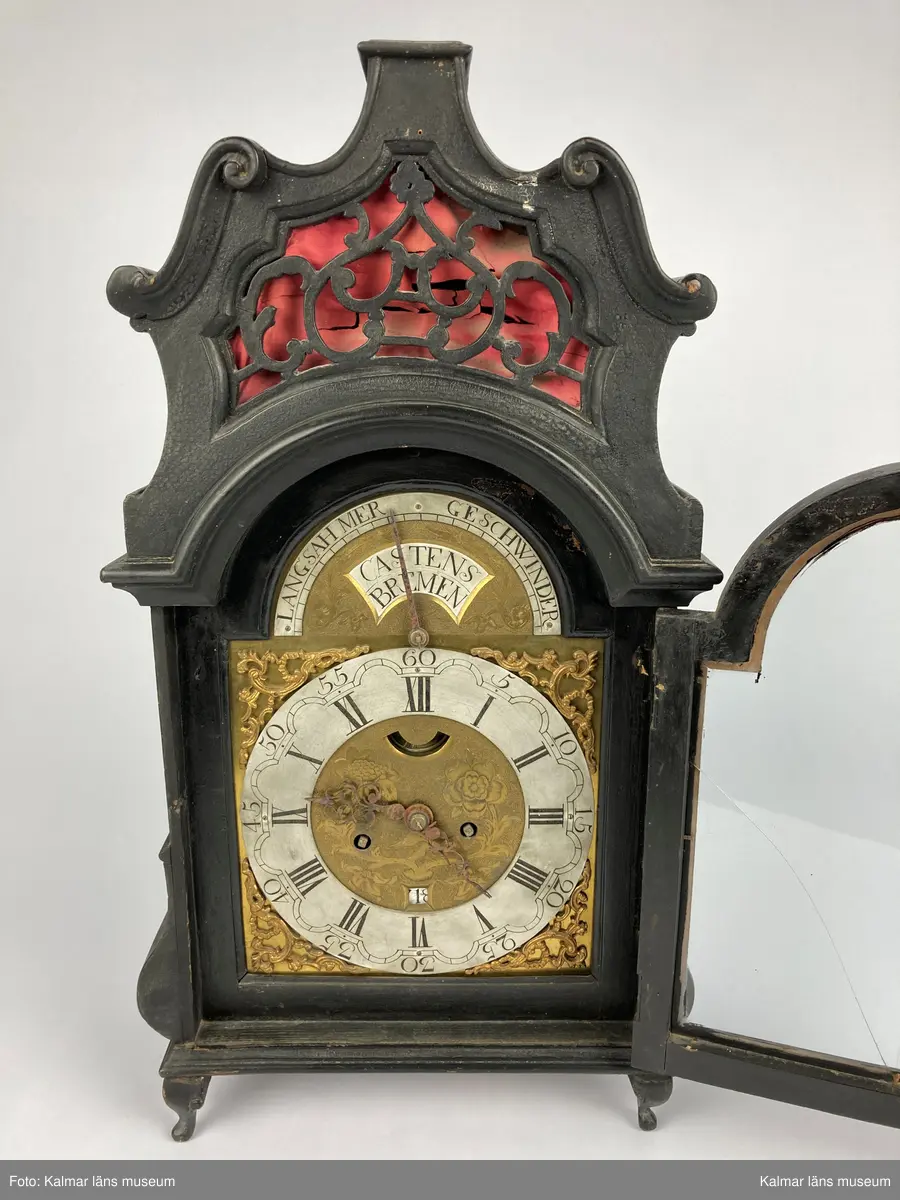 KLM 2268. Bordsur. Fodral av svartlackerat trä med genombrutna ornament med rött (blekt) siden bakom. Profilerat krön. Fodralet är på baksidan försedd med en dörr. Urverk av mässing. Bakom en glasförsedd dörr urtavlan av tenn och mässing med brännförgyllda ornament. Urtavlan är signerad: Castens Bremen. Fodralet har fyra korta profilerade ben.