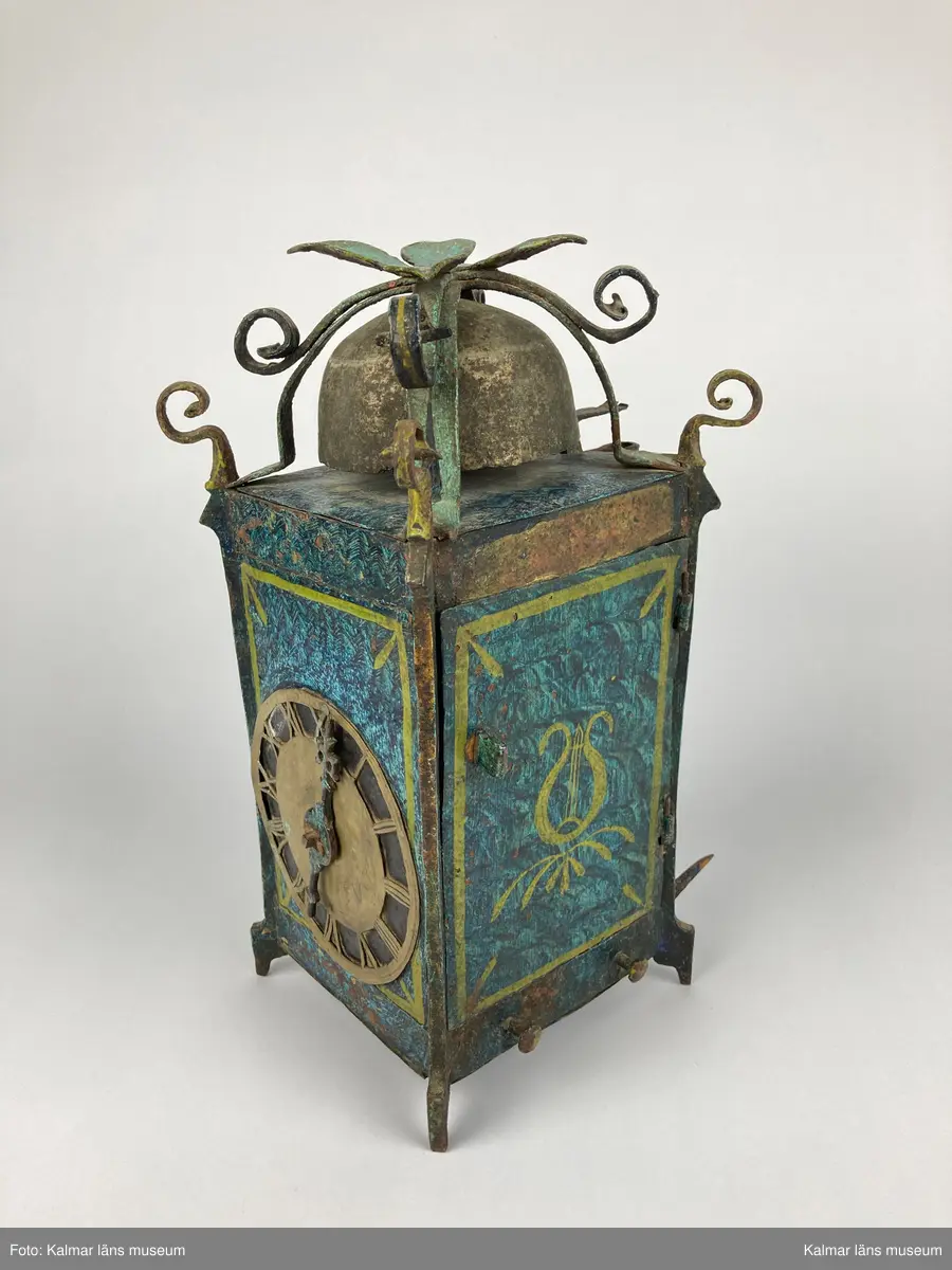 KLM 2798. Bordsur. Rektangulär stomme av järn med öppningsbara sidor. Upptill smidda detaljer samt ringklocka. På ena sidan urtavla av mässing med en visare av järn. Urverk av järn och mässing med släphaksgång. Fodralet är målat blått med gula kantlister, på sidoväggarna en lyra i gult. Uret har fyra fötter. På baksidan ögla av järn. En pendel av metall tillhör. Lodlinor, lod och trissor saknas.