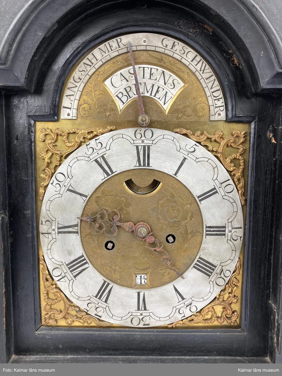 KLM 2268. Bordsur. Fodral av svartlackerat trä med genombrutna ornament med rött (blekt) siden bakom. Profilerat krön. Fodralet är på baksidan försedd med en dörr. Urverk av mässing. Bakom en glasförsedd dörr urtavlan av tenn och mässing med brännförgyllda ornament. Urtavlan är signerad: Castens Bremen. Fodralet har fyra korta profilerade ben.