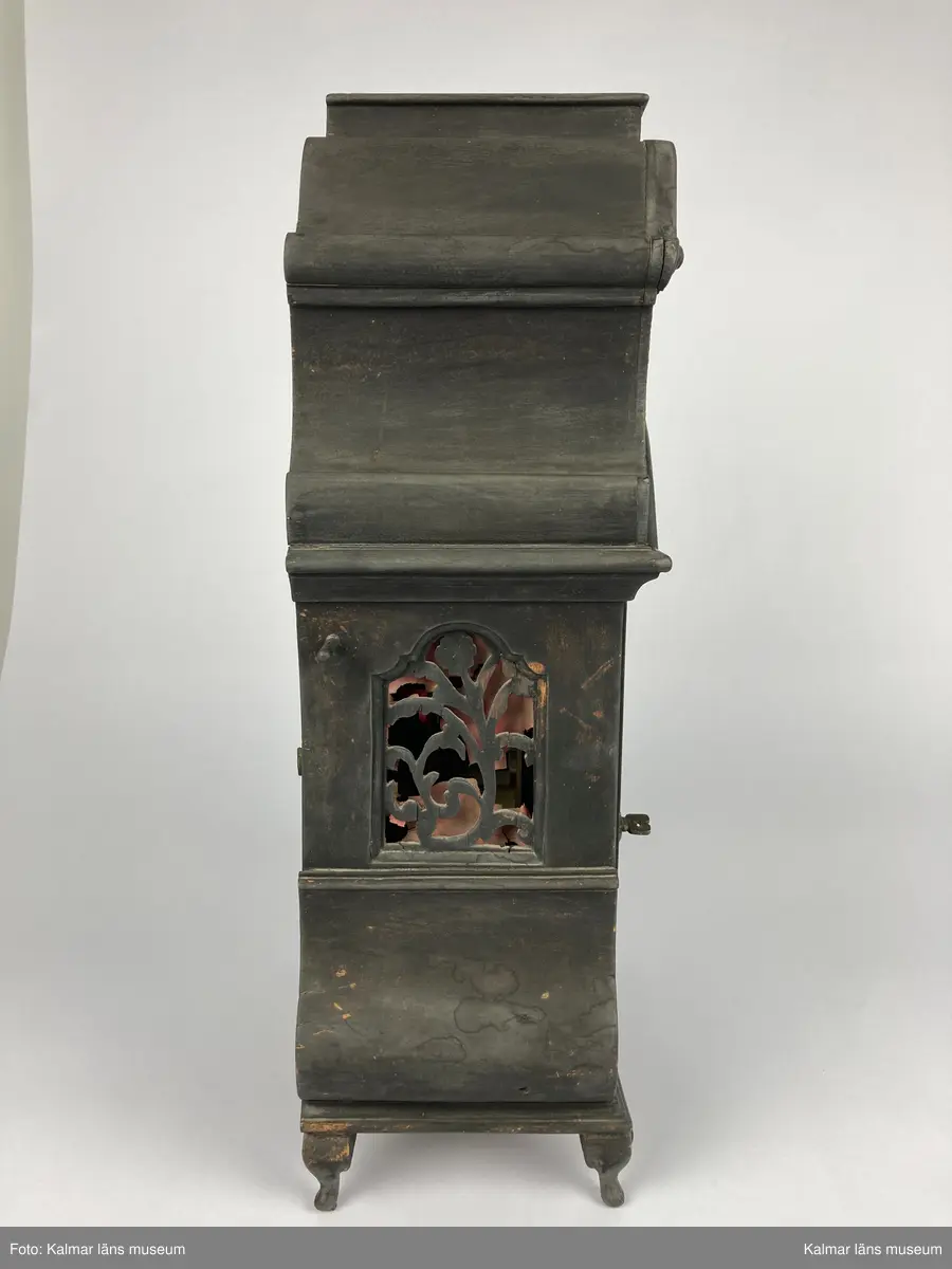 KLM 2268. Bordsur. Fodral av svartlackerat trä med genombrutna ornament med rött (blekt) siden bakom. Profilerat krön. Fodralet är på baksidan försedd med en dörr. Urverk av mässing. Bakom en glasförsedd dörr urtavlan av tenn och mässing med brännförgyllda ornament. Urtavlan är signerad: Castens Bremen. Fodralet har fyra korta profilerade ben.