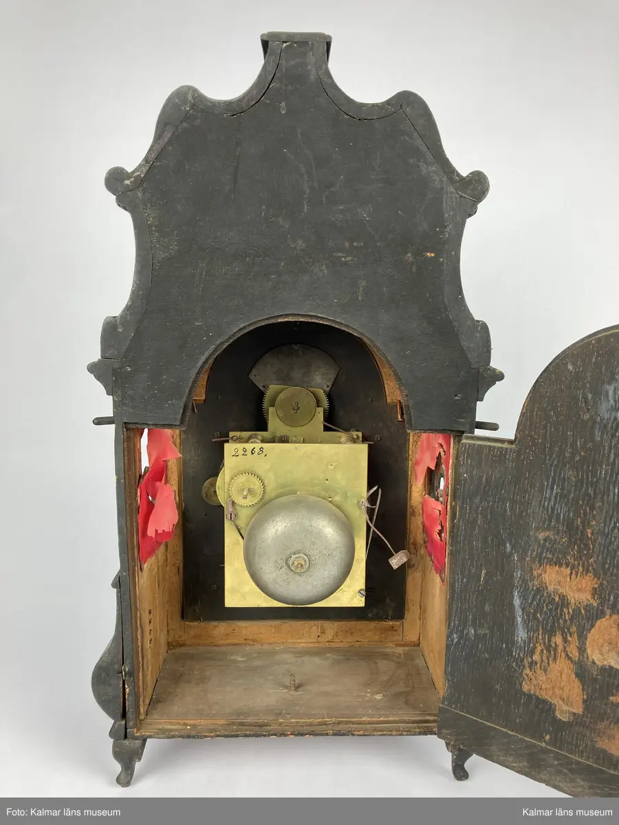 KLM 2268. Bordsur. Fodral av svartlackerat trä med genombrutna ornament med rött (blekt) siden bakom. Profilerat krön. Fodralet är på baksidan försedd med en dörr. Urverk av mässing. Bakom en glasförsedd dörr urtavlan av tenn och mässing med brännförgyllda ornament. Urtavlan är signerad: Castens Bremen. Fodralet har fyra korta profilerade ben.