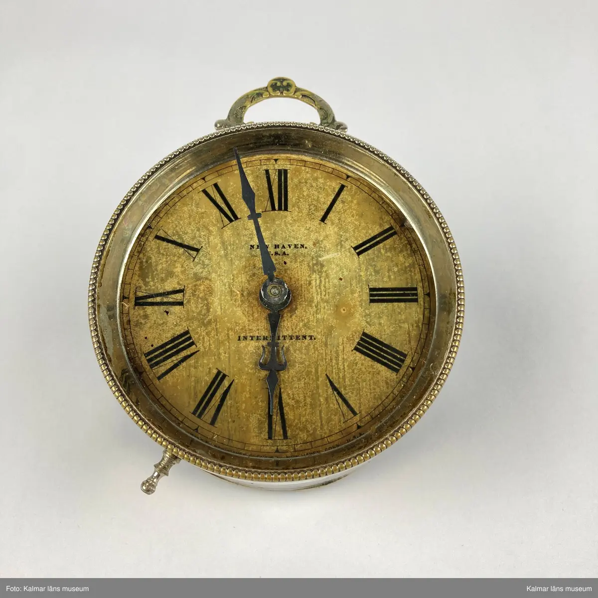 KLM 44996. Väckarklocka. Av silverfärgad metall och mässing. Inglasad urtavla med svarta visare och svarta romerska siffror. Text på urtavlan: New Haven USA Intermittent. Klockans ena ben saknas.
