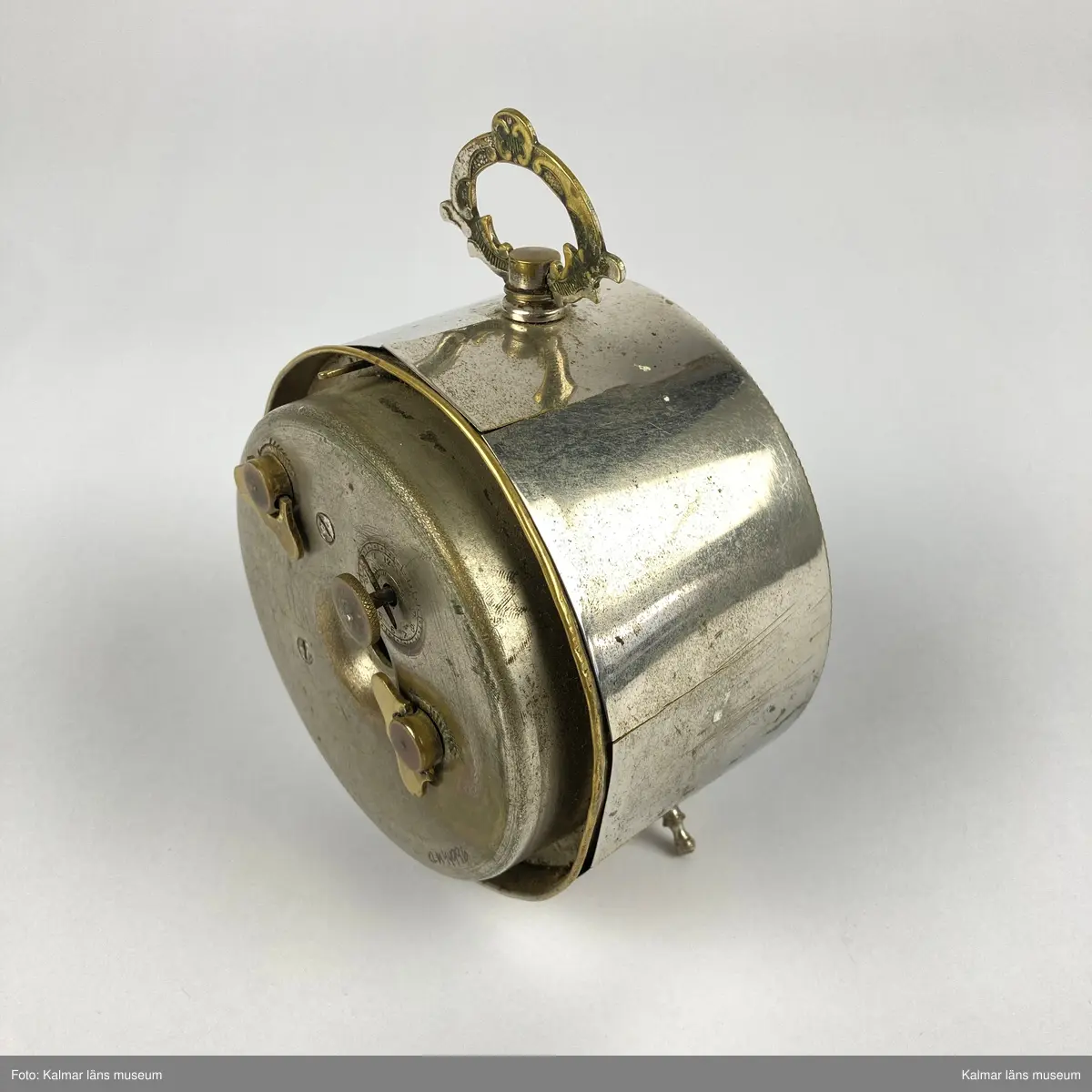 KLM 44996. Väckarklocka. Av silverfärgad metall och mässing. Inglasad urtavla med svarta visare och svarta romerska siffror. Text på urtavlan: New Haven USA Intermittent. Klockans ena ben saknas.