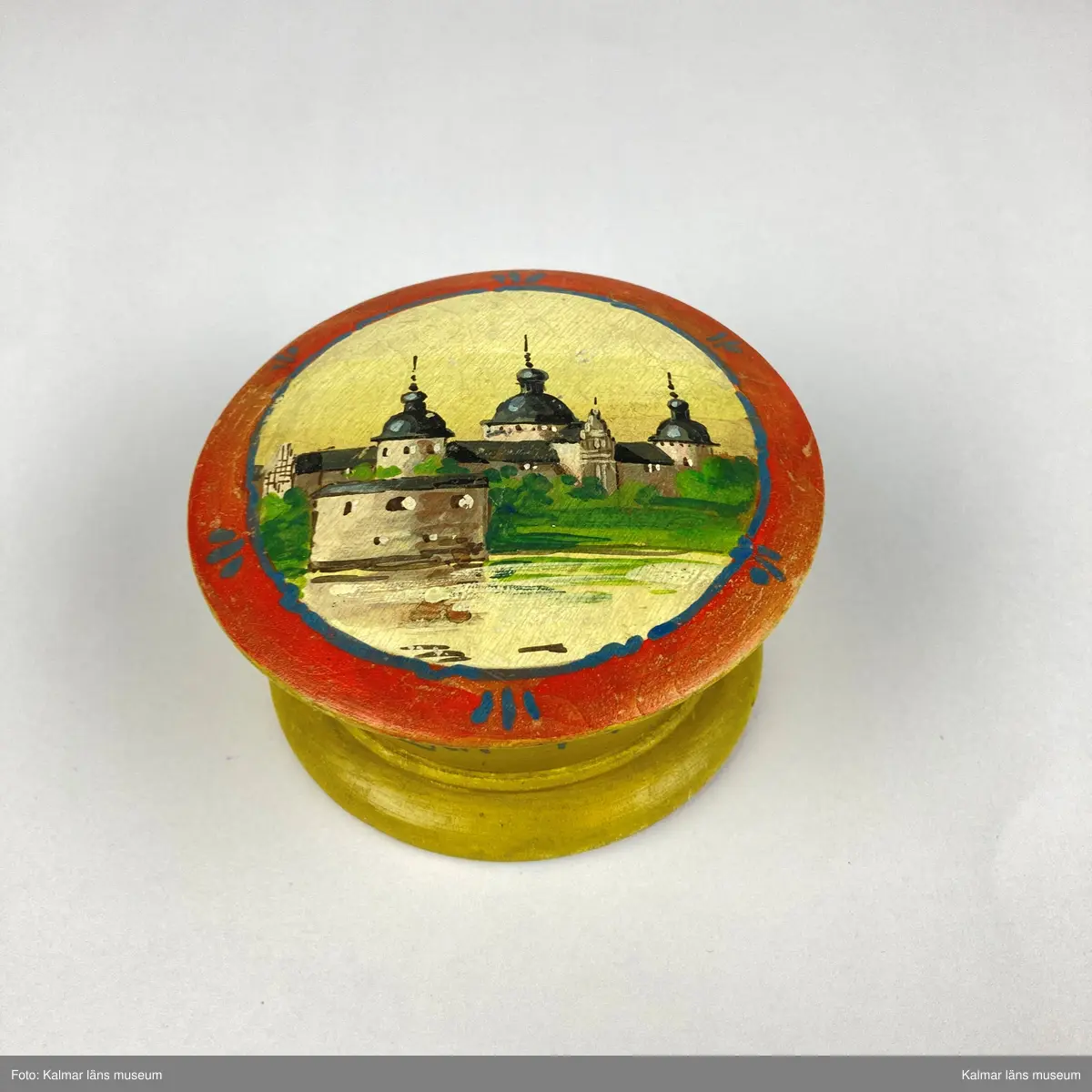 KLM 46965. Ask. Svarvad rund ask av trä med lock. Asken är målad i gult, på sidan målad text i blått: Minne Från Utställningen i KALMAR 1913. På locket målat motiv med Kalmar slott i gult, svart, grönt och grått inom en röd- och blåmålad ram. På askens undersida siffror: 875.