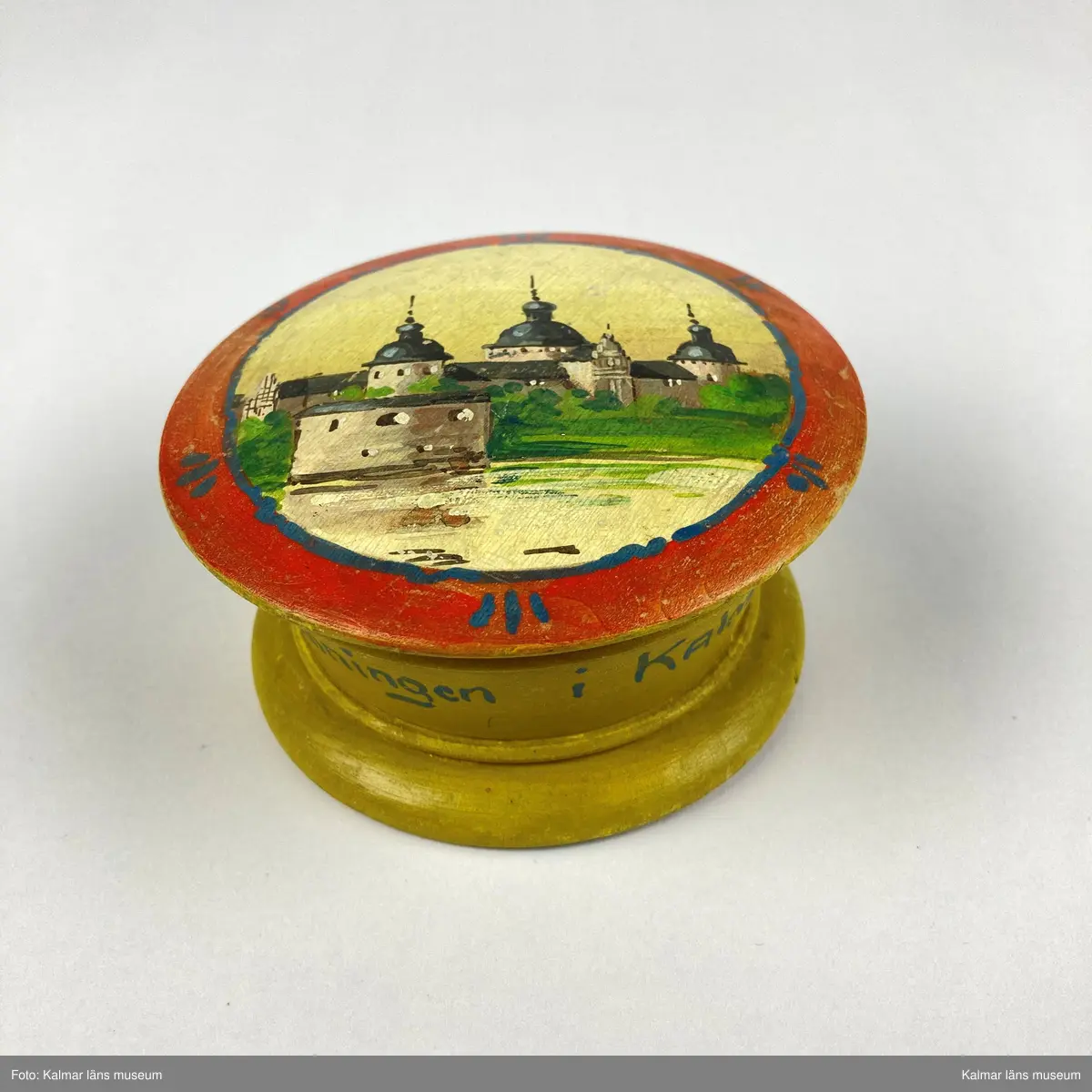 KLM 46965. Ask. Svarvad rund ask av trä med lock. Asken är målad i gult, på sidan målad text i blått: Minne Från Utställningen i KALMAR 1913. På locket målat motiv med Kalmar slott i gult, svart, grönt och grått inom en röd- och blåmålad ram. På askens undersida siffror: 875.