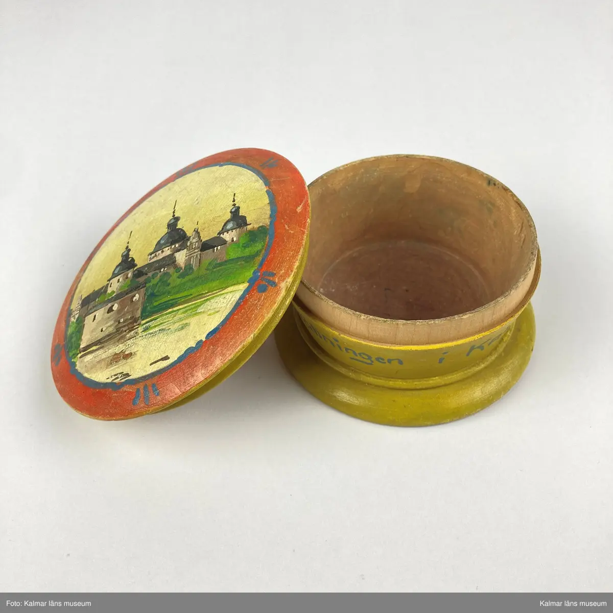 KLM 46965. Ask. Svarvad rund ask av trä med lock. Asken är målad i gult, på sidan målad text i blått: Minne Från Utställningen i KALMAR 1913. På locket målat motiv med Kalmar slott i gult, svart, grönt och grått inom en röd- och blåmålad ram. På askens undersida siffror: 875.