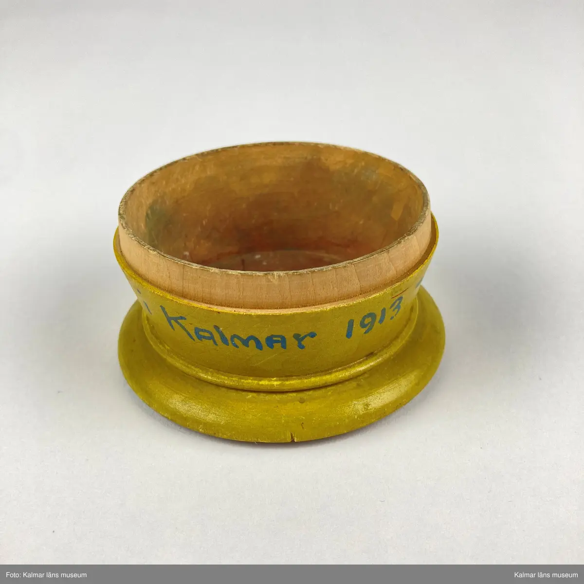 KLM 46965. Ask. Svarvad rund ask av trä med lock. Asken är målad i gult, på sidan målad text i blått: Minne Från Utställningen i KALMAR 1913. På locket målat motiv med Kalmar slott i gult, svart, grönt och grått inom en röd- och blåmålad ram. På askens undersida siffror: 875.