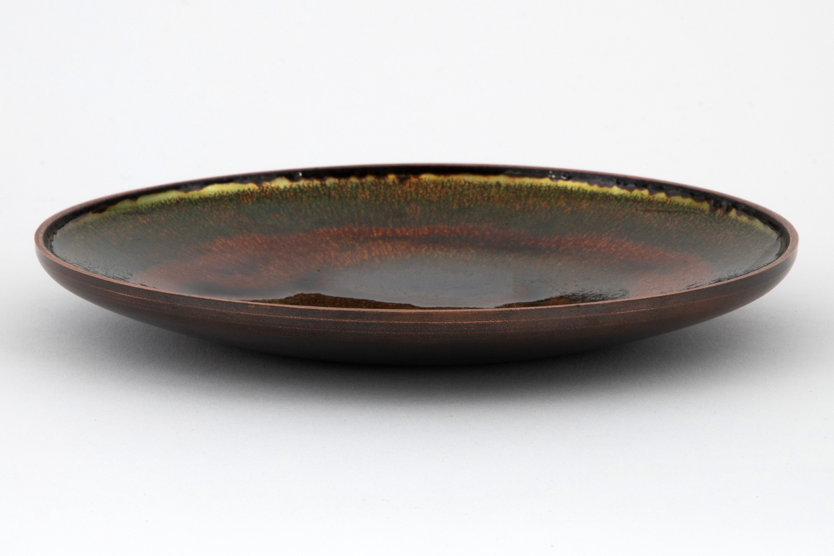 Skål i kobber med rund, konkav form. Oversiden er dekket med emalje i mosegrønt, rustrødt og brunt i forskjellige nyanser.