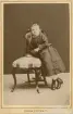 En flicka i mörk klänning lutar sig mot en stol med en bok på sitsen.
Helfigur. Ateljéfoto.
Elisabeth Eksandh (senare g. Bergstedt), Slätthög. Mormor t. givaren.