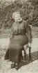 En leende kvinna i mörk blus och kjol sitter på en stol i solskenet på en gårdsplan.
Elisabeth Eksandh (senare g. Bergstedt), Slätthög. Mormor t. givaren.