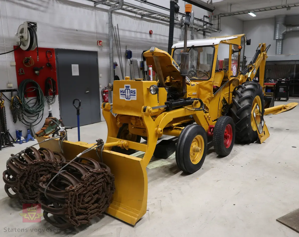 BM-Volvo T-400 traktor med Hymas type 4 graver. Gul, svart og rød traktorgraver lagd i hovedsak av metall, gummi og glass. Merket av produsentene. Dette er en jordbrukstraktor som er påmontert en graver, (gravemaskin), i bakkant. Graveren er hydraulisk drevet og har skuffe og to støtteben. Traktoren er 2-akslet har 6 hjul. To av hjulene er for å kunne bruke traktoren med halvbelter (belter følger med). Motoren er av typen D 913 fra BM, som betyr en tre sylindret firetakts diesel drevet type med slagvolum 2,5 liter. Motoren yter 47 (DIN) Hk. Motoren er bygd med basis fra en Perkins motor. Girkassen har innebygd reduksjons gir som gir åtte gir fremover og to gir bakover. Traktoren har 12 V elektrisk anlegg. Rødt sete merket med BM.
Følgende fulgte med maskinen:
- To skuffer og en hydraulikksentral samt to trekkstenger.
- Fire bilder/tegninger i glass og ramme.
- Regningsblokk.
- Innrammet avisutklipp.
