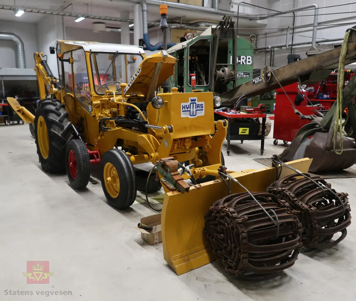 BM-Volvo T-400 traktor med Hymas type 4 graver. Gul, svart og rød traktorgraver lagd i hovedsak av metall, gummi og glass. Merket av produsentene. Dette er en jordbrukstraktor som er påmontert en graver, (gravemaskin), i bakkant. Graveren er hydraulisk drevet og har skuffe og to støtteben. Traktoren er 2-akslet har 6 hjul. To av hjulene er for å kunne bruke traktoren med halvbelter (belter følger med). Motoren er av typen D 913 fra BM, som betyr en tre sylindret firetakts diesel drevet type med slagvolum 2,5 liter. Motoren yter 47 (DIN) Hk. Motoren er bygd med basis fra en Perkins motor. Girkassen har innebygd reduksjons gir som gir åtte gir fremover og to gir bakover. Traktoren har 12 V elektrisk anlegg. Rødt sete merket med BM.
Følgende fulgte med maskinen:
- To skuffer og en hydraulikksentral samt to trekkstenger.
- Fire bilder/tegninger i glass og ramme.
- Regningsblokk.
- Innrammet avisutklipp.