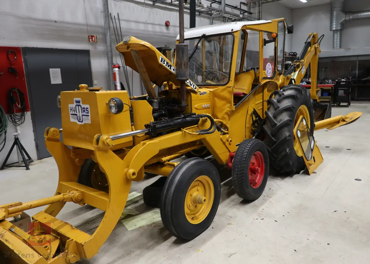 BM-Volvo T-400 traktor med Hymas type 4 graver. Gul, svart og rød traktorgraver lagd i hovedsak av metall, gummi og glass. Merket av produsentene. Dette er en jordbrukstraktor som er påmontert en graver, (gravemaskin), i bakkant. Graveren er hydraulisk drevet og har skuffe og to støtteben. Traktoren er 2-akslet har 6 hjul. To av hjulene er for å kunne bruke traktoren med halvbelter (belter følger med). Motoren er av typen D 913 fra BM, som betyr en tre sylindret firetakts diesel drevet type med slagvolum 2,5 liter. Motoren yter 47 (DIN) Hk. Motoren er bygd med basis fra en Perkins motor. Girkassen har innebygd reduksjons gir som gir åtte gir fremover og to gir bakover. Traktoren har 12 V elektrisk anlegg. Rødt sete merket med BM.
Følgende fulgte med maskinen:
- To skuffer og en hydraulikksentral samt to trekkstenger.
- Fire bilder/tegninger i glass og ramme.
- Regningsblokk.
- Innrammet avisutklipp.