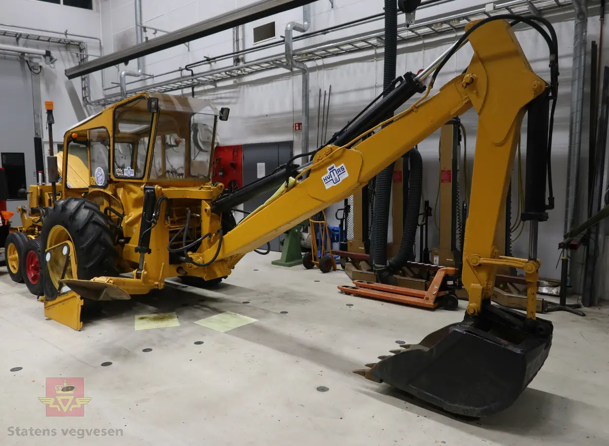 BM-Volvo T-400 traktor med Hymas type 4 graver. Gul, svart og rød traktorgraver lagd i hovedsak av metall, gummi og glass. Merket av produsentene. Dette er en jordbrukstraktor som er påmontert en graver, (gravemaskin), i bakkant. Graveren er hydraulisk drevet og har skuffe og to støtteben. Traktoren er 2-akslet har 6 hjul. To av hjulene er for å kunne bruke traktoren med halvbelter (belter følger med). Motoren er av typen D 913 fra BM, som betyr en tre sylindret firetakts diesel drevet type med slagvolum 2,5 liter. Motoren yter 47 (DIN) Hk. Motoren er bygd med basis fra en Perkins motor. Girkassen har innebygd reduksjons gir som gir åtte gir fremover og to gir bakover. Traktoren har 12 V elektrisk anlegg. Rødt sete merket med BM.
Følgende fulgte med maskinen:
- To skuffer og en hydraulikksentral samt to trekkstenger.
- Fire bilder/tegninger i glass og ramme.
- Regningsblokk.
- Innrammet avisutklipp.