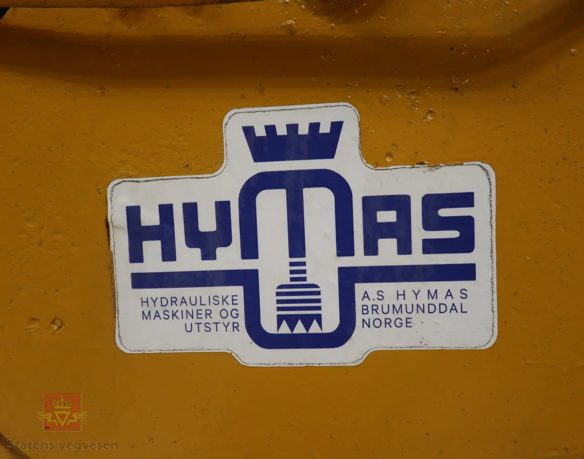 BM-Volvo T-400 traktor med Hymas type 4 graver. Gul, svart og rød traktorgraver lagd i hovedsak av metall, gummi og glass. Merket av produsentene. Dette er en jordbrukstraktor som er påmontert en graver, (gravemaskin), i bakkant. Graveren er hydraulisk drevet og har skuffe og to støtteben. Traktoren er 2-akslet har 6 hjul. To av hjulene er for å kunne bruke traktoren med halvbelter (belter følger med). Motoren er av typen D 913 fra BM, som betyr en tre sylindret firetakts diesel drevet type med slagvolum 2,5 liter. Motoren yter 47 (DIN) Hk. Motoren er bygd med basis fra en Perkins motor. Girkassen har innebygd reduksjons gir som gir åtte gir fremover og to gir bakover. Traktoren har 12 V elektrisk anlegg. Rødt sete merket med BM.
Følgende fulgte med maskinen:
- To skuffer og en hydraulikksentral samt to trekkstenger.
- Fire bilder/tegninger i glass og ramme.
- Regningsblokk.
- Innrammet avisutklipp.