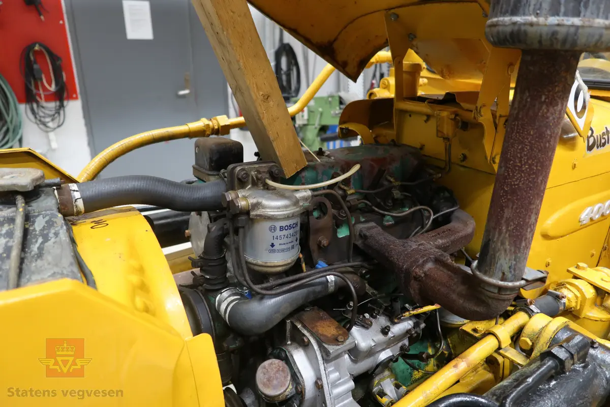 BM-Volvo T-400 traktor med Hymas type 4 graver. Gul, svart og rød traktorgraver lagd i hovedsak av metall, gummi og glass. Merket av produsentene. Dette er en jordbrukstraktor som er påmontert en graver, (gravemaskin), i bakkant. Graveren er hydraulisk drevet og har skuffe og to støtteben. Traktoren er 2-akslet har 6 hjul. To av hjulene er for å kunne bruke traktoren med halvbelter (belter følger med). Motoren er av typen D 913 fra BM, som betyr en tre sylindret firetakts diesel drevet type med slagvolum 2,5 liter. Motoren yter 47 (DIN) Hk. Motoren er bygd med basis fra en Perkins motor. Girkassen har innebygd reduksjons gir som gir åtte gir fremover og to gir bakover. Traktoren har 12 V elektrisk anlegg. Rødt sete merket med BM.
Følgende fulgte med maskinen:
- To skuffer og en hydraulikksentral samt to trekkstenger.
- Fire bilder/tegninger i glass og ramme.
- Regningsblokk.
- Innrammet avisutklipp.