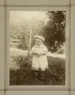 En liten flicka i tvärrandig klänning med invävt mönster och ljus sjömansmössa poserar för fotografen i en trädgård.
Margareta Bergstedt (senare g. Lidén), Växjö.