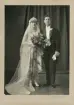 Ett brudpar hos fotografen. Bruden i knäkort vit brudklänning med slöja, brudgummen i frack med handskar.
Helfigur. Ateljéfoto.
Under bilden står antecknat: 
