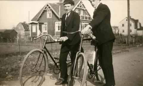 Två unga män står med sina cyklar på en grusig landsväg. Lägg märke till det stora huset i bakgrunden med snickarglädje under takåsarna. Båda männen bär skärmmössor och mörka kostymer.